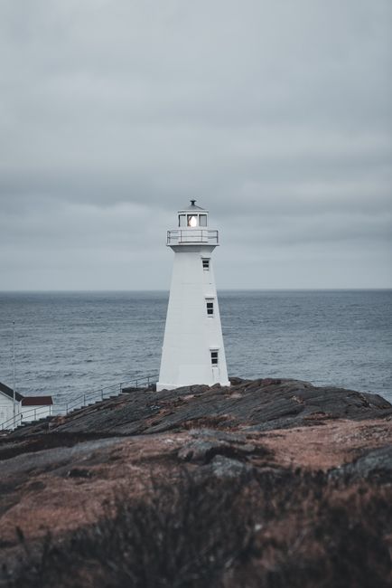 A tall, white lighthouse with a cylindrical body and a small balcony near the top, standing on a rocky shoreline overlooking the sea. The lighthouse features several small, rectangular windows and a l