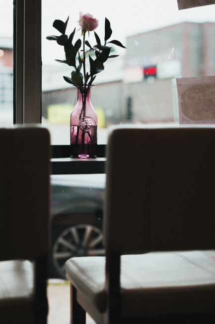 Inside a home during a house removal process, a narrow view shows two dark upholstered chairs in the foreground, partially obstructing the scene. Behind them, a window provides natural light and reveals an urban street with a blurred vehicle passing by. On the windowsill, there is a pink glass vase containing a single pink rose with green leaves, positioned centrally. The interior environment appears clean and organized, with soft lighting highlighting the flowers and the window's view. This setting may be during packing or preparing for furniture transport, consistent with professional house removals offered by Man with Van St Johns Wood. The overall scene reflects a moment in home relocation or packing, emphasizing the careful handling of personal items and the logistical process of furniture and belongings transport.