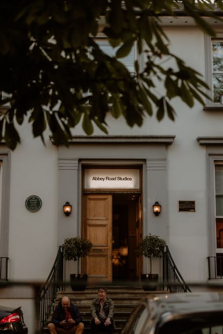 The exterior of a building associated with Abbey Road Studios, with a white facade and a wooden double door entrance illuminated by two small wall-mounted lamps. The entrance is accessed by a short flight of steps with black metal railings on either side, and two potted plants are positioned on the landings near the door. In the foreground, two men are seated on the steps, one appears to be using a mobile device while the other is engaged in conversation, with a van parked nearby. The scene is partially framed by overhanging tree branches and leaves at the top of the image, indicating an outdoor setting during daylight hours. This image, associated with house removals and furniture transport, reflects a moving process near a well-known music studio in St Johns Wood, as displayed on the sign above the doorway.